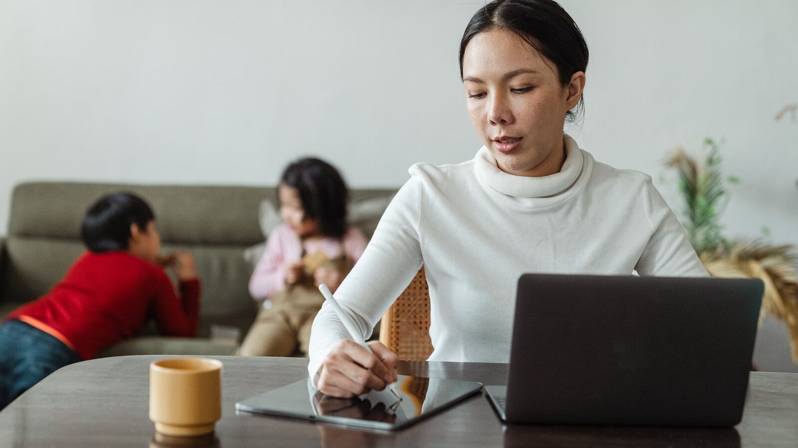 Focused young Asian woman in casual wear writing with stylus on tablet and browsing laptop while working remotely at home with children