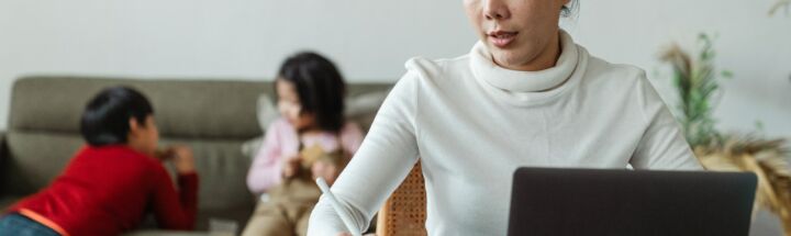 Focused young Asian woman in casual wear writing with stylus on tablet and browsing laptop while working remotely at home with children