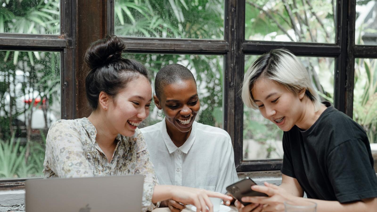 Cheerful young diverse female friends laughing while watching funny video on smartphone during coffee break in cozy cafe