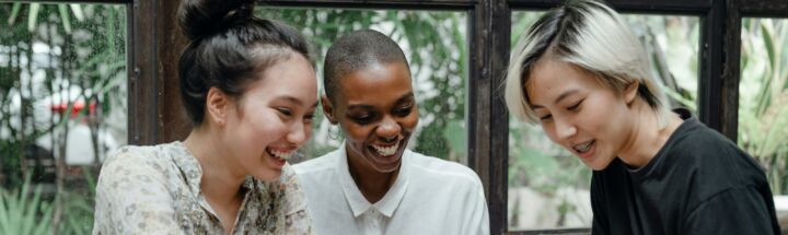 Cheerful young diverse female friends laughing while watching funny video on smartphone during coffee break in cozy cafe