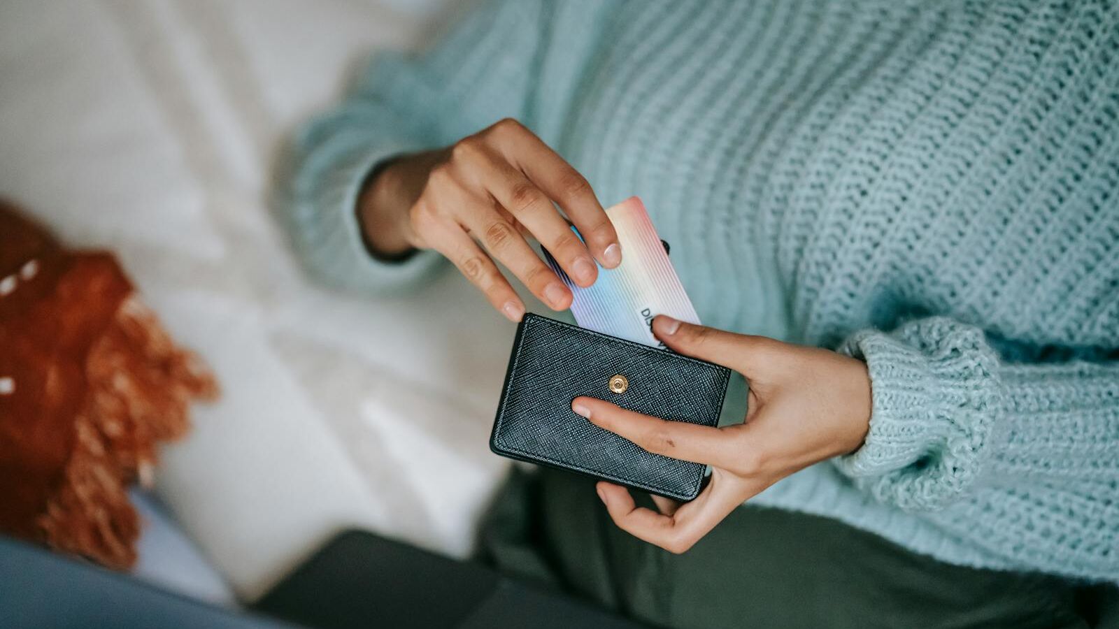 From above crop anonymous female in casual sweater taking credit card from wallet and lying on bed with laptop before making online purchase
