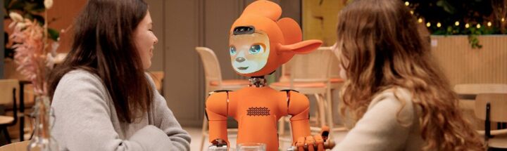 Two women talk to an orange robot at a table.