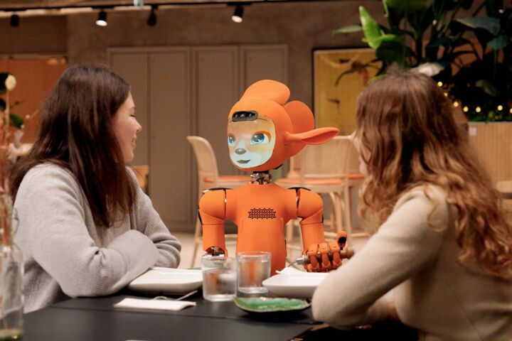 Two women talk to an orange robot at a table.
