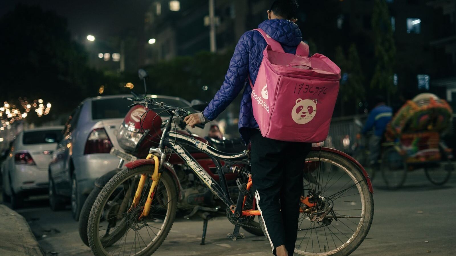 A bicycle delivery person walks on a city street at night with a food delivery backpack.